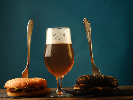A traditional and black burger on a wooden table and with glasses full of foamy light beer.