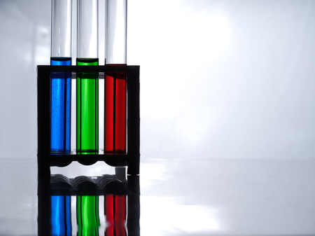 Test tubes with blue, green and red liquid in a rack for test tubes on a white table and a white...