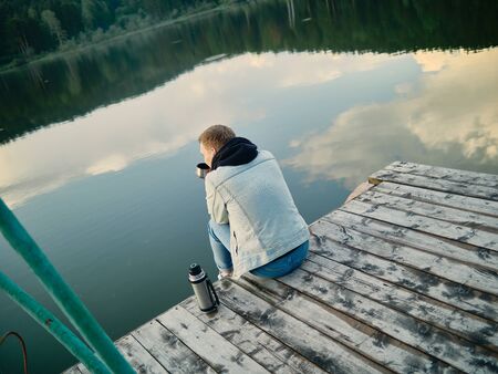 Rest on the lake. A young guy in blue jeans, a denim jacket and a black hoodie is sitting on a wooden pier against the background of the lake and the forest with a flask in his hands.の写真素材