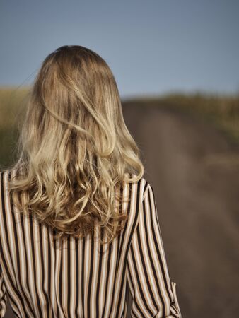 The blonde is walking along a country road in a striped dress from the backの写真素材
