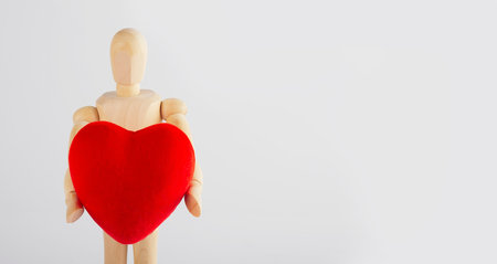 Wooden man holding a heart figurine in his hands on white background. Wooden mannequin. Valentine's day concept.の写真素材