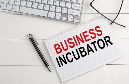 BUSINESS INCUBATOR text on notebook with keyboard , pen glasses on a white wooden backgroundの写真素材