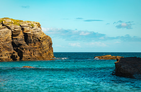 Rocky coastal cliff with turquoise waters and distant rocks under clear blue sky.の写真素材
