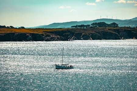 Boat floating on shimmering water with a rocky coastline and distant wind turbines under clear sky.の写真素材