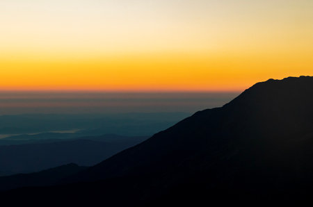 Sunrise over mountain silhouettes with a vibrant gradient sky transitioning from dark orange to pale yellow, creating serene and tranquil atmosphere.の写真素材