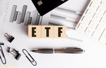Wooden blocks spelling ETF on a financial desk with graphs, calculator, credit card and office...