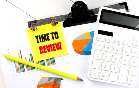 Sticky note with phrase time to review attached to financial documents with a pie charts, bar graphs, calculator and yellow pencil.の写真素材