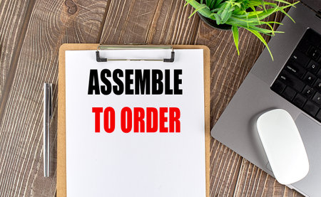 Assemble to order phrase on clipboard with a laptop, pen and plant on wooden desk representing...
