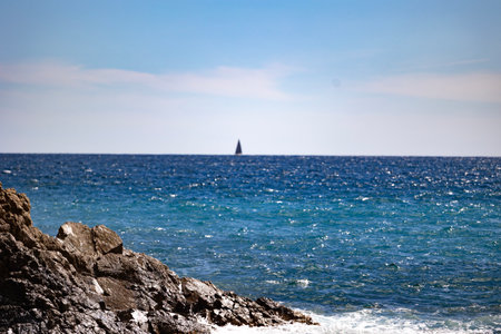 Rocky coastline with a deep blue ocean and a distant sailboat on the horizon under a clear sky.の写真素材