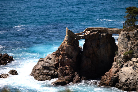 Stone bridge connecting rocky cliffs over turquoise ocean waves with a dramatic coastal scenery.