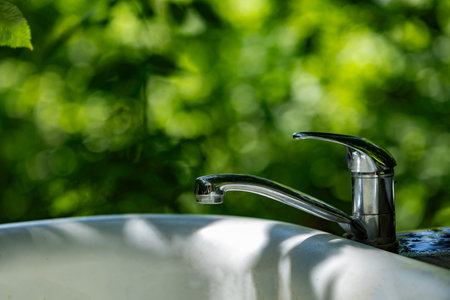 Metal faucet on white sink outdoors with sunlight and green forest background in soft focus.の写真素材