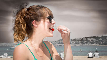 Close-up of a pretty, young, red-headed woman eating a cherry. She is at the beach and Emits happiness, joy of life and pleasure.の写真素材