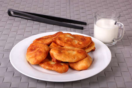 Fried cakes on white plate, kitchen tongs and cup of milk on gray wicker backgroundの写真素材