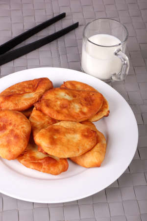 Fried cakes on white plate, kitchen tongs and cup of milk on gray wicker background. Vertical photoの写真素材
