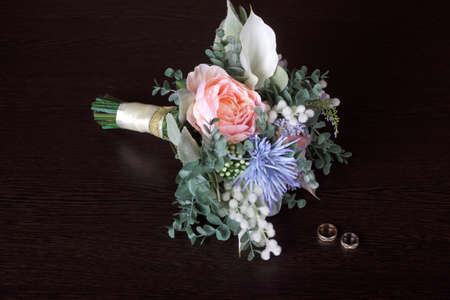 Wedding bouquet of beautiful artificial flowers and wedding rings on a dark table in low lightの写真素材