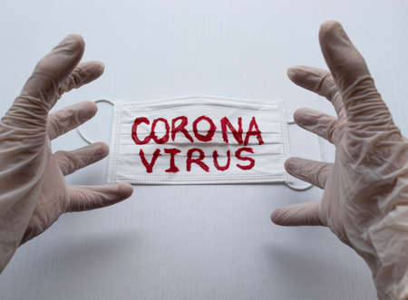 Closed up to White Esterilized gloves making an angry hand expression over white face mask with red CORONAVIRUS inscription with white blackground. COVID-2019の写真素材