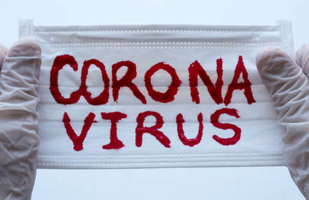 Closed up to White Face Mask with red CORONAVIRUS inscription and two hands with White Gloves or Esterilized globes holding them. COVID-2019の写真素材