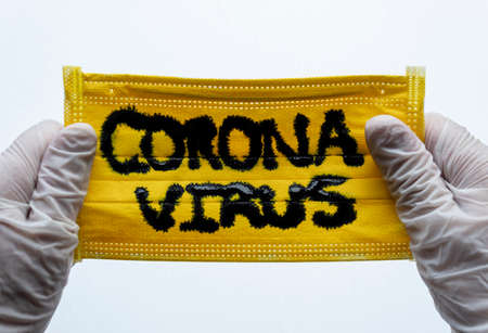 Closed up to Yellow Face Mask with CORONAVIRUS inscription and two hands with White Gloves or Esterilized globes holding them. COVID-2019の写真素材