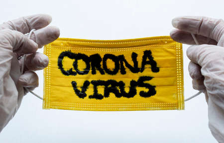 Closed up to Yellow Face Mask with CORONAVIRUS inscription and two hands with White Gloves or Esterilized globes holding them. COVID-2019の写真素材