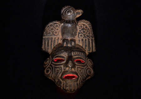 An ancient ceramic pre columbus mask based in American indigenous tribes art illuminated by red light inside and white light over black backgroundの写真素材