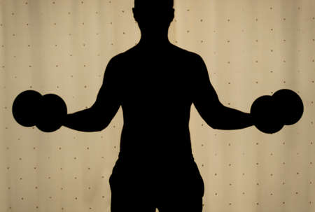Silhouette of a young man making a biceps dumbbell curl with yellow curtains at background. Ideal for fitness, sports, home workout, Indoor exercise and healthy life.の写真素材