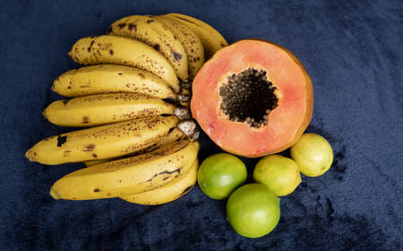 Top view of a fresh sliced orange papaya with seeds inside, a freckled bananas cluster and three green lemons over a blue navy and black background. Fruit and eating concept photographyの写真素材