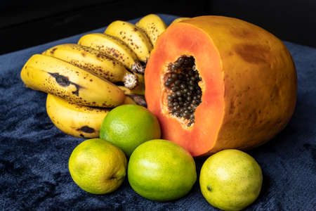 Close up to a fresh sliced orange papaya with seeds inside, a freckled bananas cluster and three green lemons over a blue navy and black background. Fruit and eating concept photographyの写真素材