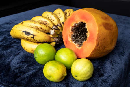 Close up to a fresh sliced orange papaya with seeds inside, a freckled bananas cluster and three green lemons over a blue navy and black background. Fruit and eating concept photographyの写真素材