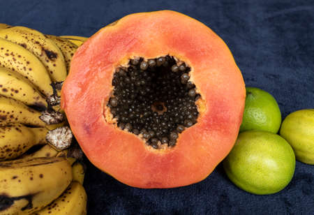 Close up to a top view of a fresh sliced orange papaya with seeds inside, a freckled bananas cluster and three green lemons over a blue navy background. Fruit and eating concept photographyの写真素材