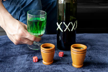 Close up to a man holding a cup of green homemade liquor with two wooden cups and green artesanal alcohol bottle and double six red dices. Party and liquor games conceptの写真素材