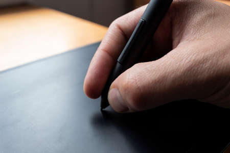 Close up to a male hand holding up a black digital pencil over a black and green drawing tablet in desk and home office background. Ideal for graphic design, web and every relationated with technologyの写真素材