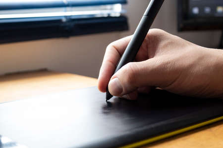Close up to a male hand holding up a black digital pencil over a black and green drawing tablet in desk and home office background. Ideal for graphic design, web and every relationated with technologyの写真素材
