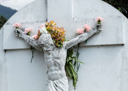 Crucified White JesusChrist Stone into a Cemetery adorned with multicolor flowers in hands, arms and flowers like a crown in head.のeditorial素材
