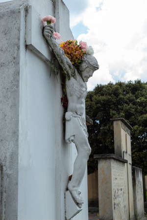 Crucified White Jesus Christ Stone into a Cemetery adorned with multicolor flowers in hands, arms and flowers like a crown in head. Big tree and cloudy sky at backgroundのeditorial素材