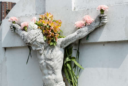 Crucified White JesusChrist Stone into a Cemetery adorned with multicolor flowers in hands, arms and flowers like a crown in head.のeditorial素材