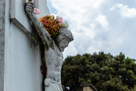 Crucified White Jesus Christ Stone into a Cemetery adorned with multicolor flowers in hands, arms and flowers like a crown in head. Big tree and cloudy sky at backgroundのeditorial素材