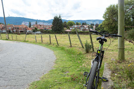 A black mountain bike at curve road over green grass and near to a barbed wire and country side and rural houses with city at backgroundの写真素材