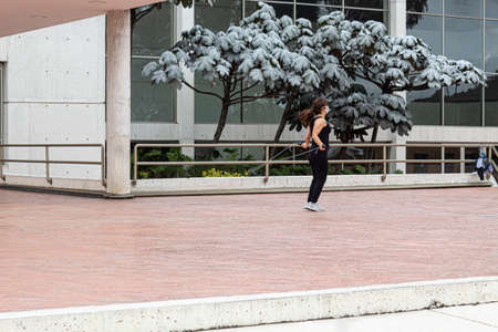 A mature woman wearing a facemask doing street workout jumping rope over a brick floor with green trees and modern architecture at background.の写真素材