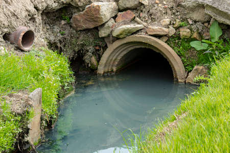 Closeup to a storm drain outlet with a contaminated river and green grassの写真素材