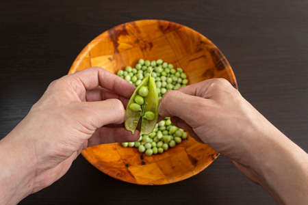 Closeup to a male hands threshing fresh green peas and a wooden bowl with green peas over a dark wooden table.の写真素材