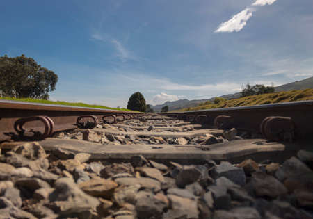 Ground view of an old railroad crossing a green country field with trees, blue sky and mountains at background in sunny morning.の写真素材