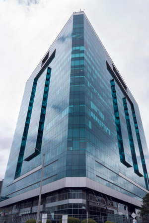 Low angle view of a triangular modern blue window skyscraper reflecting gray cloudy sky