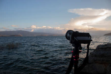Close up to a modern camera over a tripod with blue lake, mountains and orange sunset scene at backgroundの写真素材