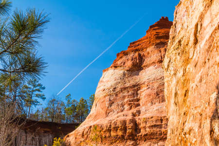 Red loamy wall of Providence Canyon with trees on the background of clear sky with contrail, USA