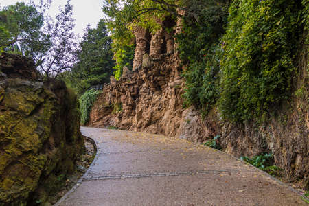 A footpath between two stone walls with lush vegetation in the Park Guell in overcast autumn day, Barcelona, Catalonia, Spainの写真素材