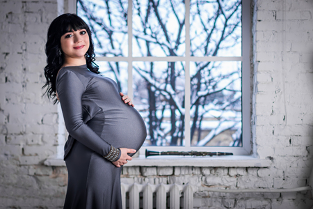 Pregnant woman stands at the window in the gray dress, winter landscape outsideの写真素材