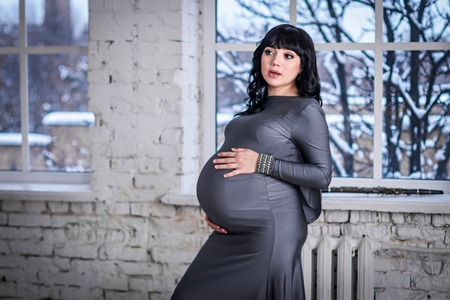 Pregnant woman stands at the window in the gray dress, winter landscape outsideの写真素材