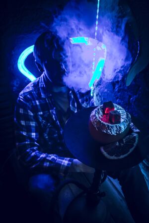 young guy smokes hookah in neon light barの写真素材
