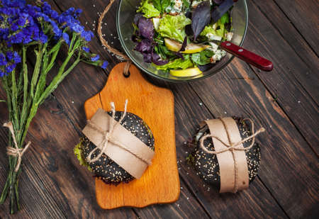 Delicious black burgers in craft paper and blue flowers on dark wood table.