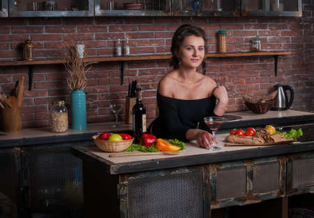 Beautiful woman with red wine glass sitting in kitchen with vegetables on tableの写真素材